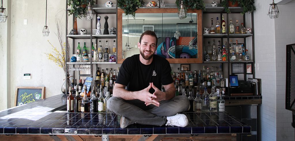 Fordham graduate Matt Trebek sits on the bar at Oso, the Mexican restaurant he opened in 2016 in the Harlem's Hamilton Heights neighborhood.