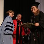 A girl wearing a black graduation gown smiles and receives a plaque from a woman wearing a light blue graduation gown