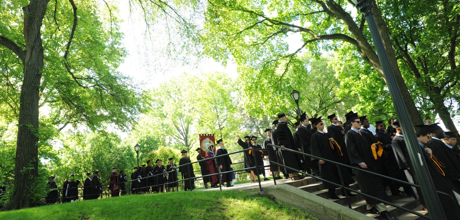 Students wearing black graduation gowns walking in a line in front of a backdrop of green foliage