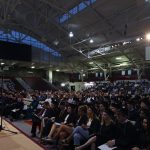 A girl wearing a black graduation gown speaks to an audience of hundreds of people