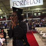 A girl wearing a black graduation gown holds hands to her tear-streaked face and walks along an aisle