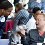 A man bends down to speak to another man seated at a table