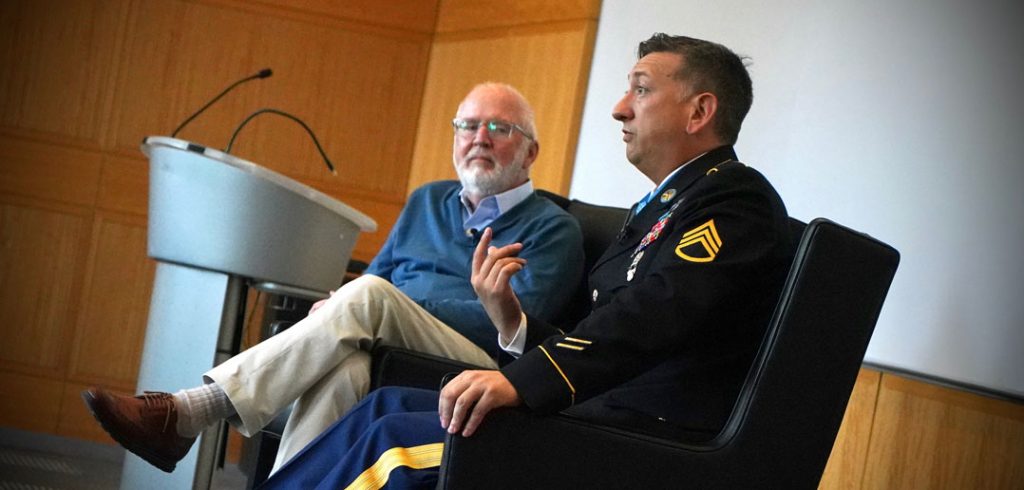 A man in a military uniform speaks next to a man wearing a blue sweater.