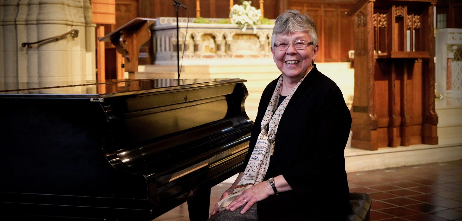 An elderly woman sits next to a piano in a church and smiles.