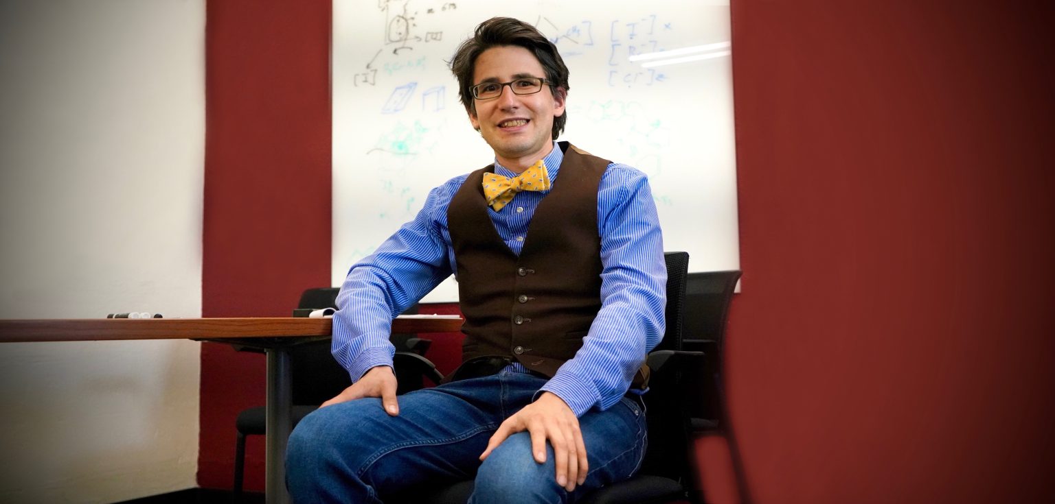 A man wearing a bow tie smiles in a classrooms setting.