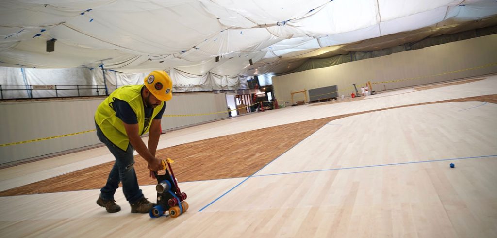 A construction worker moves machinery across a new wooden basketball court floor.