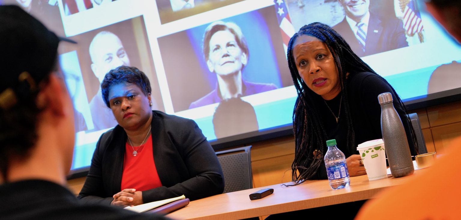 Two women speaking in front of a PowerPoint presentation featuring photos of politicians.