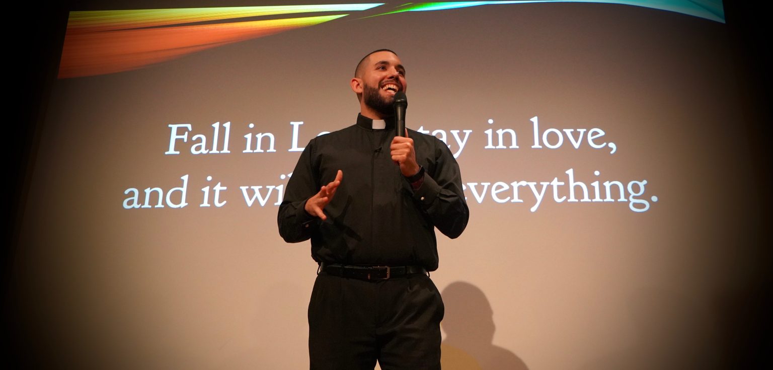 A Jesuit scholastic wearing black garb speaks in front of a PowerPoint presentation slide.