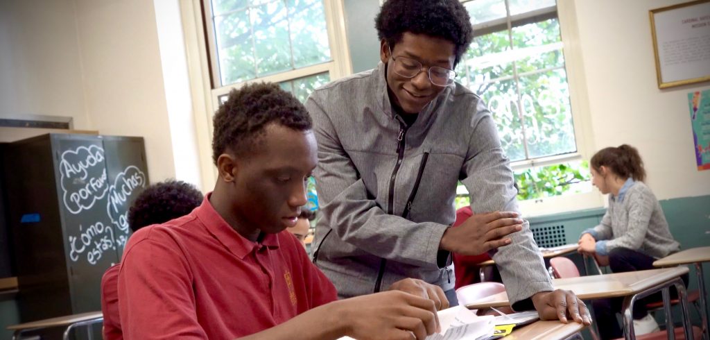 A student stands above a seated student, helping him with his studies.