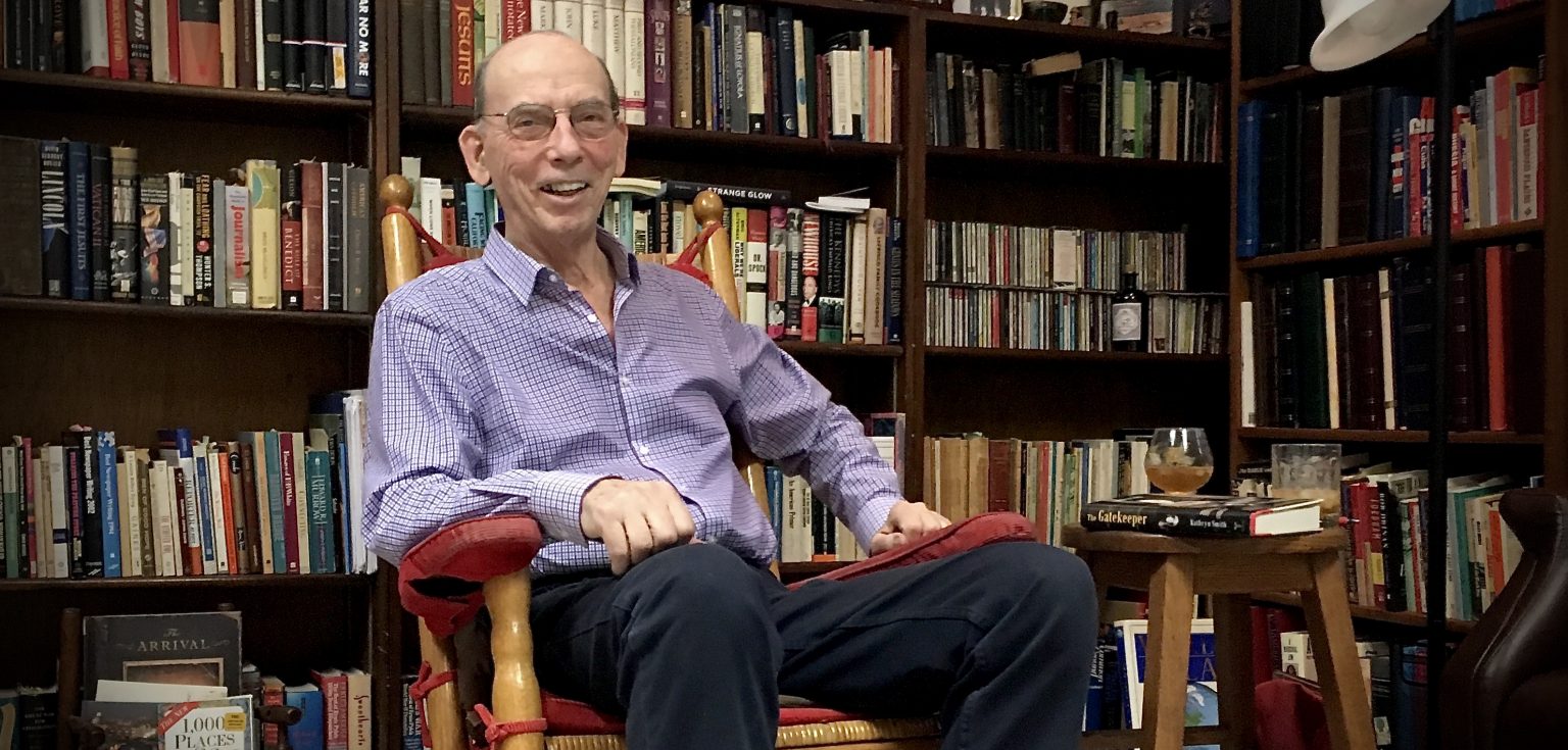 A smiling elderly man sitting in front of a row of bookshelves