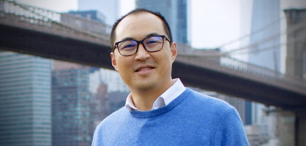 A man wearing black glasses and a blue sweater smiles in front of a bridge.