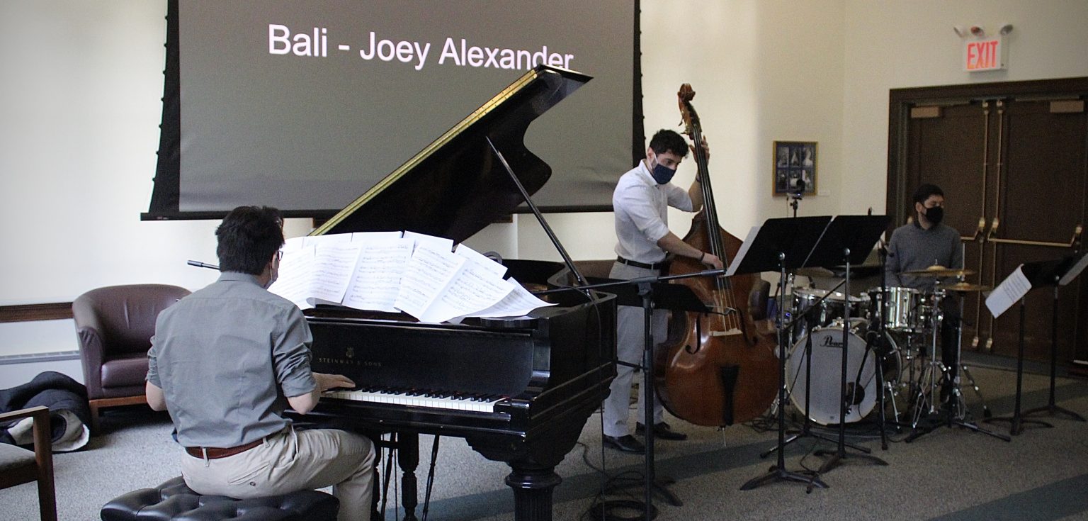 Three men each play an instrument: a piano, a cello, and a drum set, in a room with white walls.
