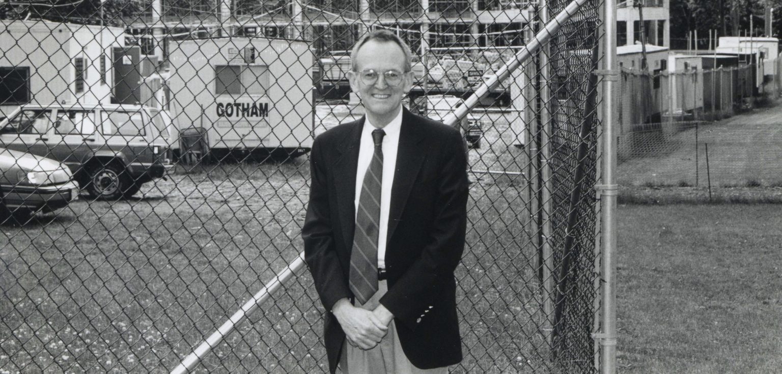A black and white photo of an elderly man smiling in front of a construction site
