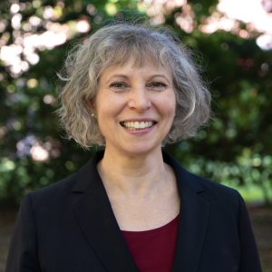 A woman with gray hair smiles in front of a green leafy background.