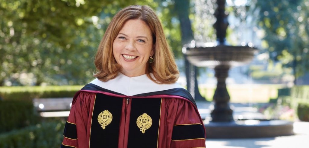 A woman with red hair and maroon graduation robes smiles in front of an outdoor fountain.