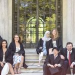 A group of Fordham graduates in cap and gown pose on the steps of Cunniffe House, with its white columns and galss doors