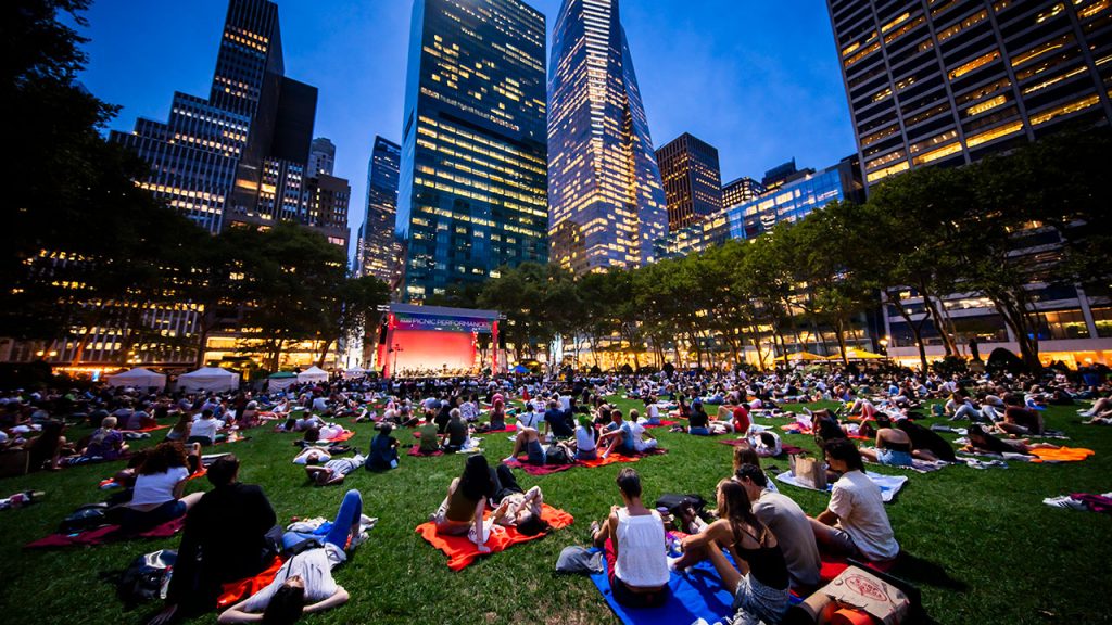 People sitting at Bryant Park grass at night with blankets, looking at stage.