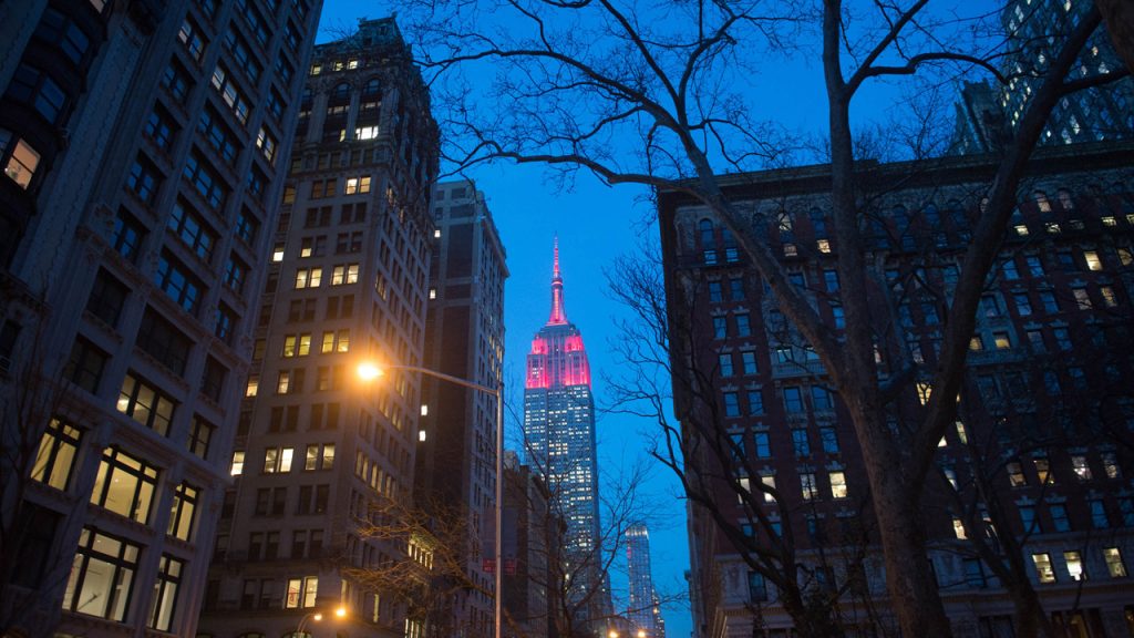 Empire State Building lit up maroon. Buildings in forefront. View is from the ground looking up.