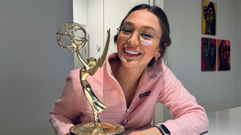 A woman smiles while posing behind an Emmy statuette.