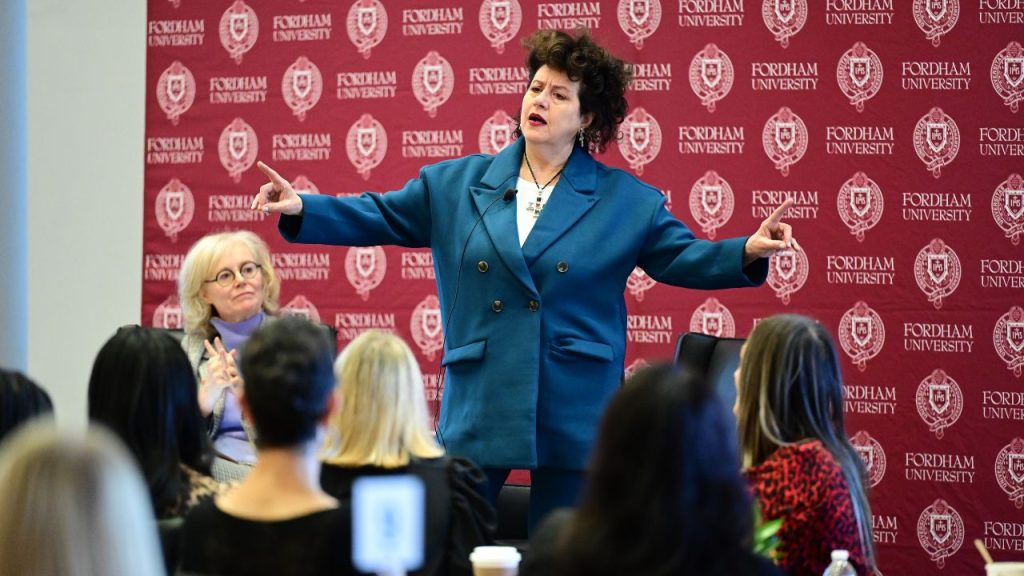 A woman in a blue blazer addresses a crowd of women in front of a background that says "Fordham." Adriana Trigiani