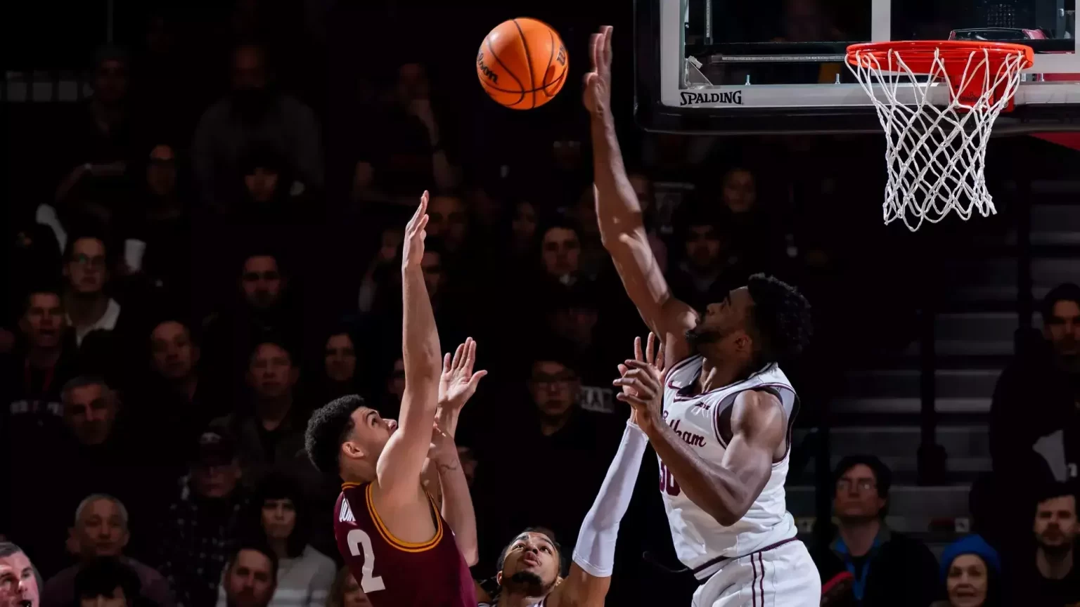 Two men playing basketball on opposing teams, jumping in air to shoot and block a basket.