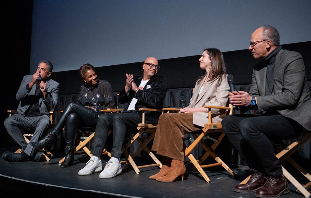 Five people sit in folding chairs on a stage, the bottom of a movie screen visible behind them