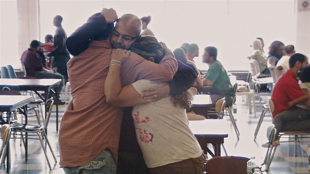 Three people embrace standing in a room where other people are seated in conversation in the background