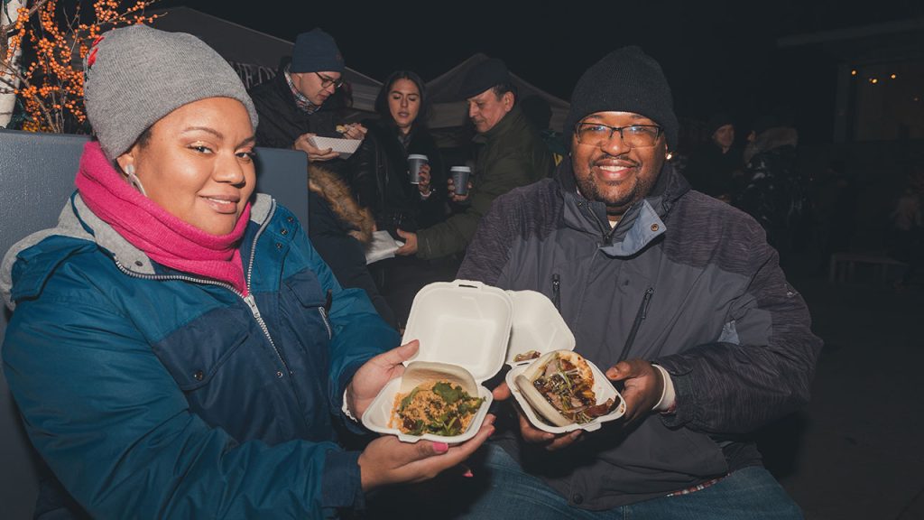 Man and woman sitting next to each other showing food in take out containers.