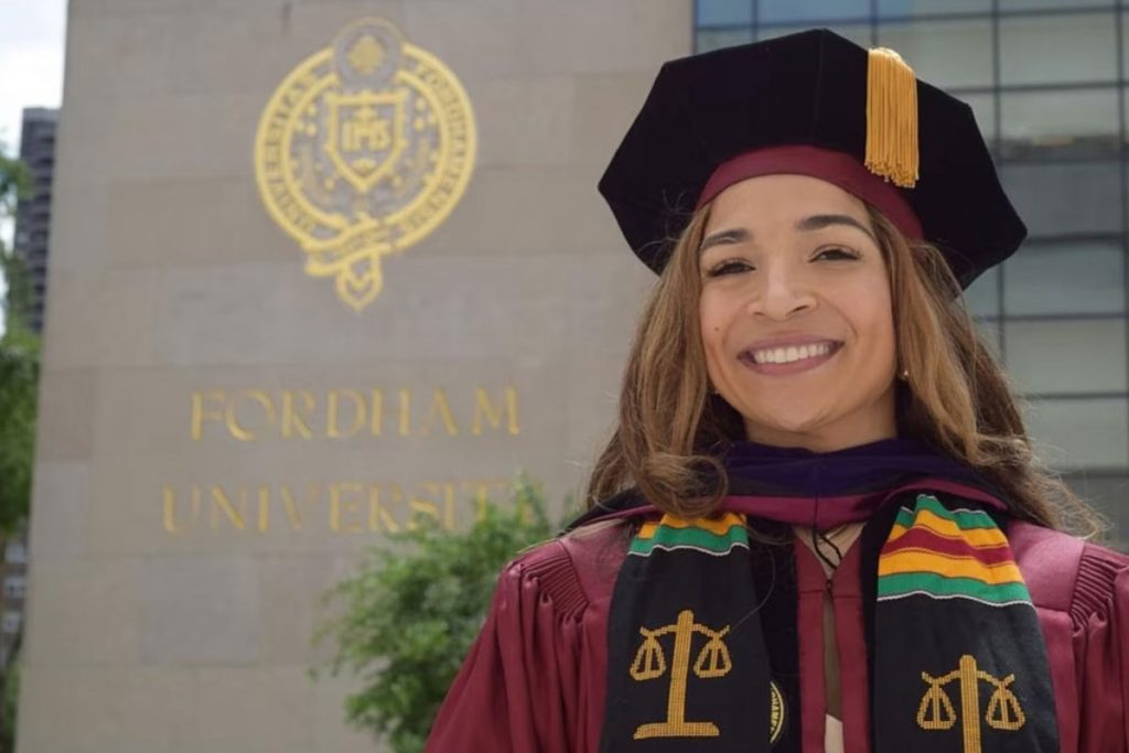 Afrika Owes smiles in her Fordham Law School graduation cap and gown in front of a Fordham building at the University's Lincoln Center campus in Manhattan