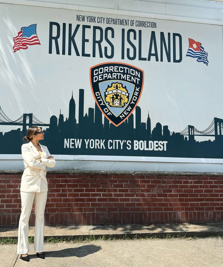 Afrika Owes, arms folded, wearing a white pantsuit, looks up at a sign that reads in part "New York City Department of Correction, Rikers Island, New York City's Boldest," and shows a silhouette of the NYC skyline.