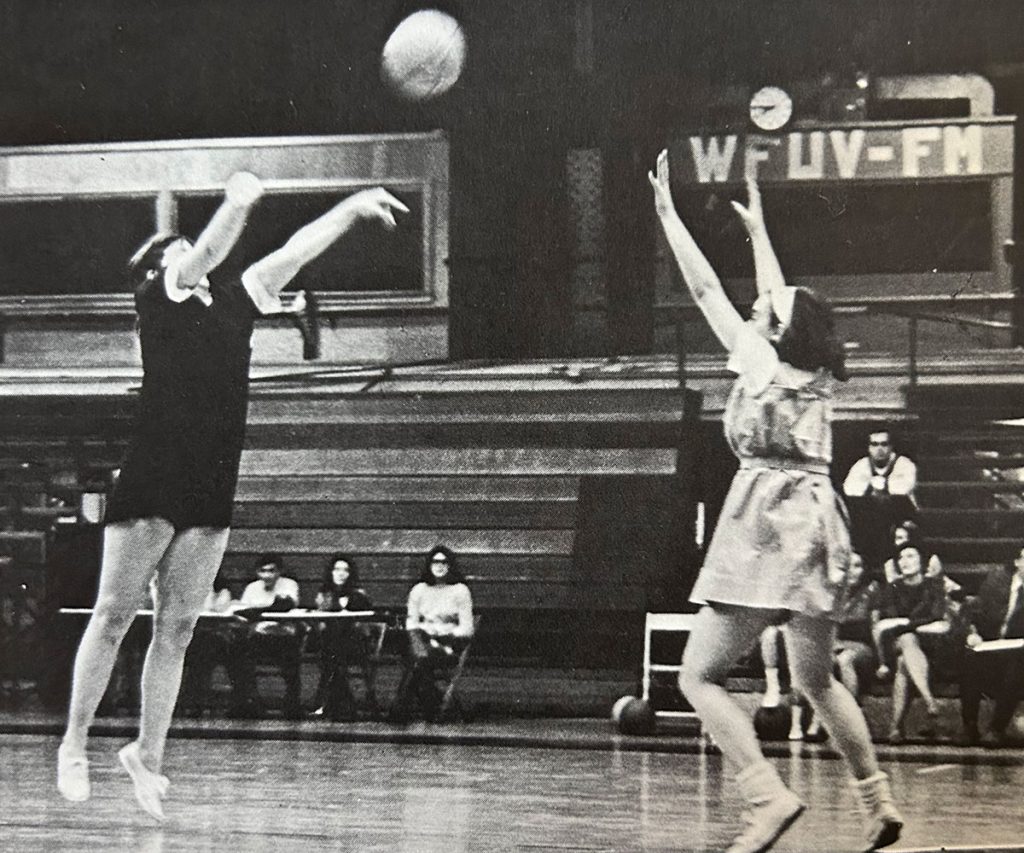 A Fordham women's basketball player releases a shot above the outstretched hands of a defender in the Rose Hill Gym, a WFUV-FM sign visible in the background