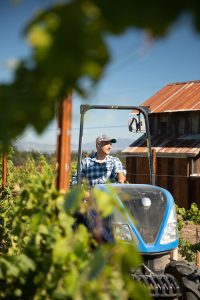 Winemaker Jamie Kutch drives a battery-operated tractor in a vineyard, vines visible in the foreground.