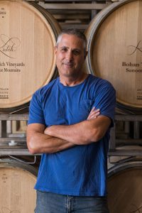 Winemaker Jamie Kutch, in blue T-shirt and jeans, smiles at camera with arms folded in front of wine barrels