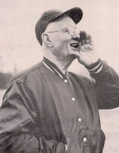 Longtime Fordham coach and athletic director Jack Coffey in Fordham hat and jacket calls out to someone off camera, left hand cupped by his mouth. The text reads Jack Coffey Day, May 17th, 1958, Fordham University