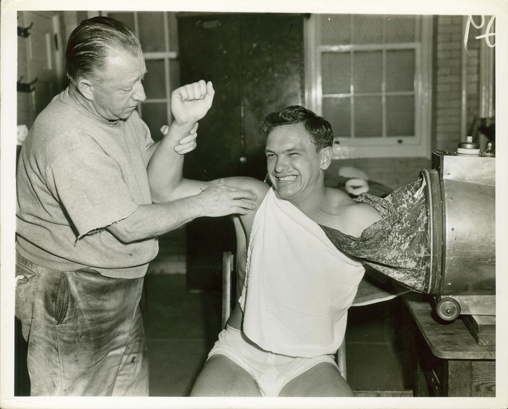 An athletic trainer holds the arm and massages the shoulder of an athlete sitting in a chair and wincing and smiling as his other arm is inside a metal device