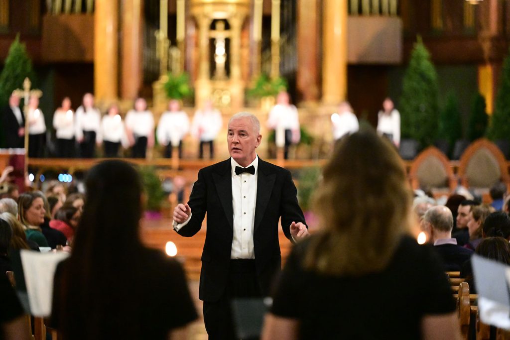 A man wearing a tuxedo conducting students in song. 