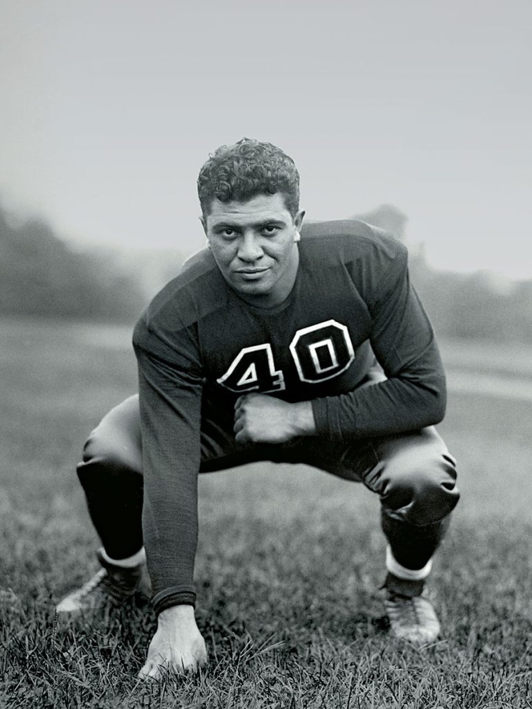 Vince Lombardi in his No. 40 Fordham uniform looks at the camera as he crouches in a football stance, one fist on the grass