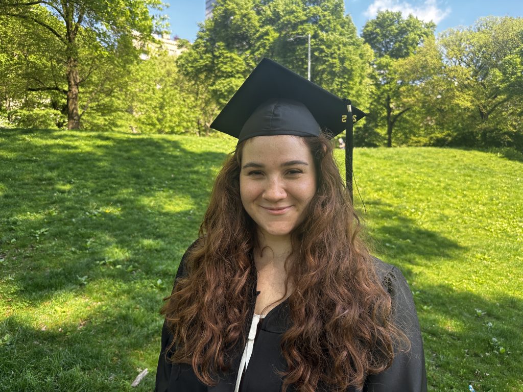 Woman smiling in cap and gown in greenery. Hills and trees behind her.