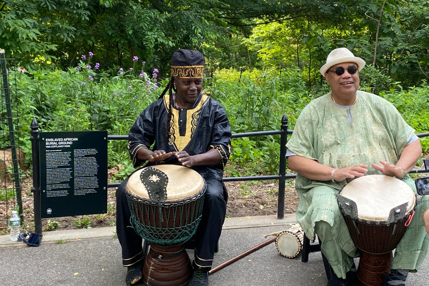 Two men sitting, playing drumbs outside.