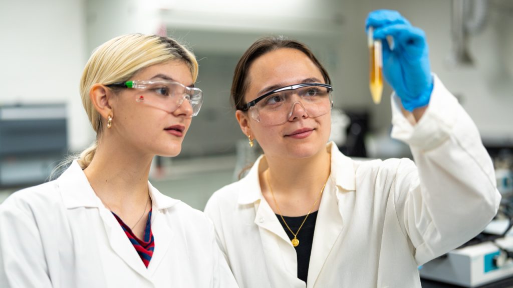 Fordham students Liana Cutter and Alexandra Ren in lab coats and safety glasses examining a test tube