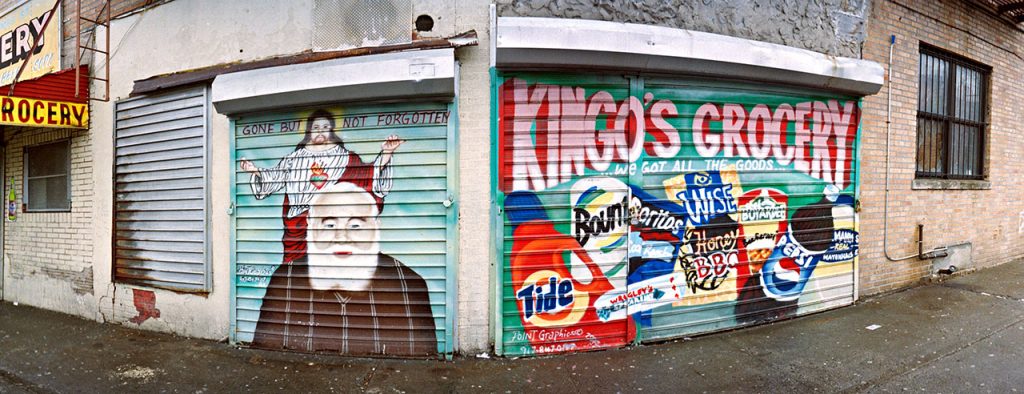 A panoramic image of a street corner storefront with multiple metal security gates. The main gate on the right is painted with a colorful advertisement for 'KINGO'S GROCERY - WE GOT ALL THE GOODS,' featuring large painted logos of common consumer products like Tide, Budweiser, and Pepsi. The adjacent gate on the left is painted with a memorial mural. This mural features a portrait of a man with a white beard wearing a brown shirt, with Jesus Christ painted behind him, arms outstretched. Text above them reads, 'GONE BUT NOT FORGOTTEN.' A red and yellow 'GROCERY' sign hangs over an unpainted section of the store on the far left.