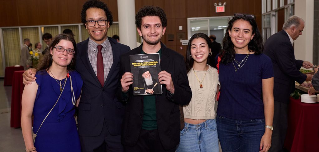 Five people stand together smiling for the camera, while one holds a flyer advertising the appearance of His All-Holiness Bartholomew, Archbishop of Constantinople-New Rome and Ecumenical Patriarch
