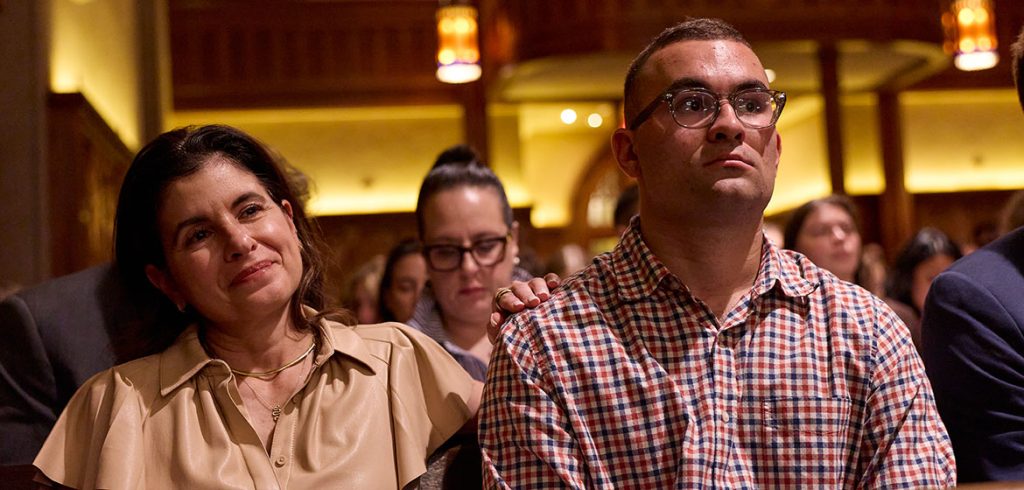 A woman wearing beige places her hand on the shoulder a man wearing a checkered shirt and glasses.