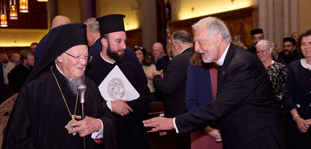 A man in black with a white collar reaches out to His All-Holiness Bartholomew, Archbishop of Constantinople-New Rome and Ecumenical Patriarch, to shake his hand.