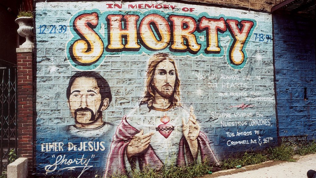 A memorial mural painted on a brick wall in the Bronx. The mural is dedicated 'IN MEMORY OF SHORTY' in large red and gold bubble letters. Below this, there are two painted portraits: one of a man with a mustache and goatee, identified as 'ELMER DE JESUS 'Shorty',' and one of Jesus Christ with a Sacred Heart. The dates '12-21-39' and '7-13-94' flank the top title. Text on the right side reads, 'YOU WILL ALWAYS LIVE IN OUR HEARTS' and 'SIEMPRE VIVIRAS EN NUESTROS CORAZONES' with an address that appears to be 'Cromwell Ave & 167th'."