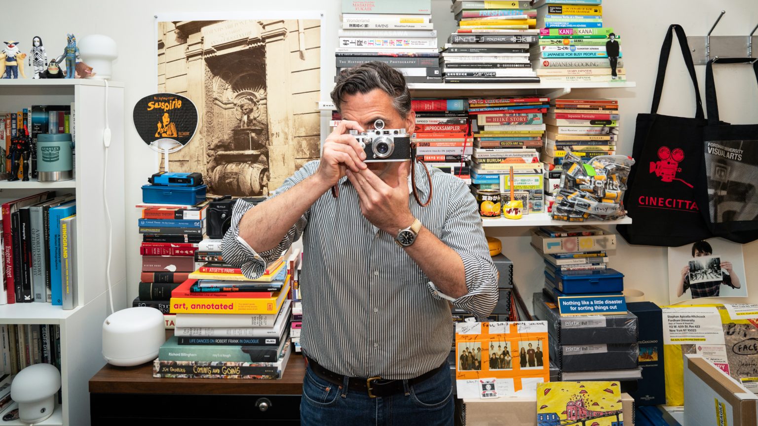 Stephan Apicella-Hitchcock holds a camera to his face and pretends to take a picture in his office.