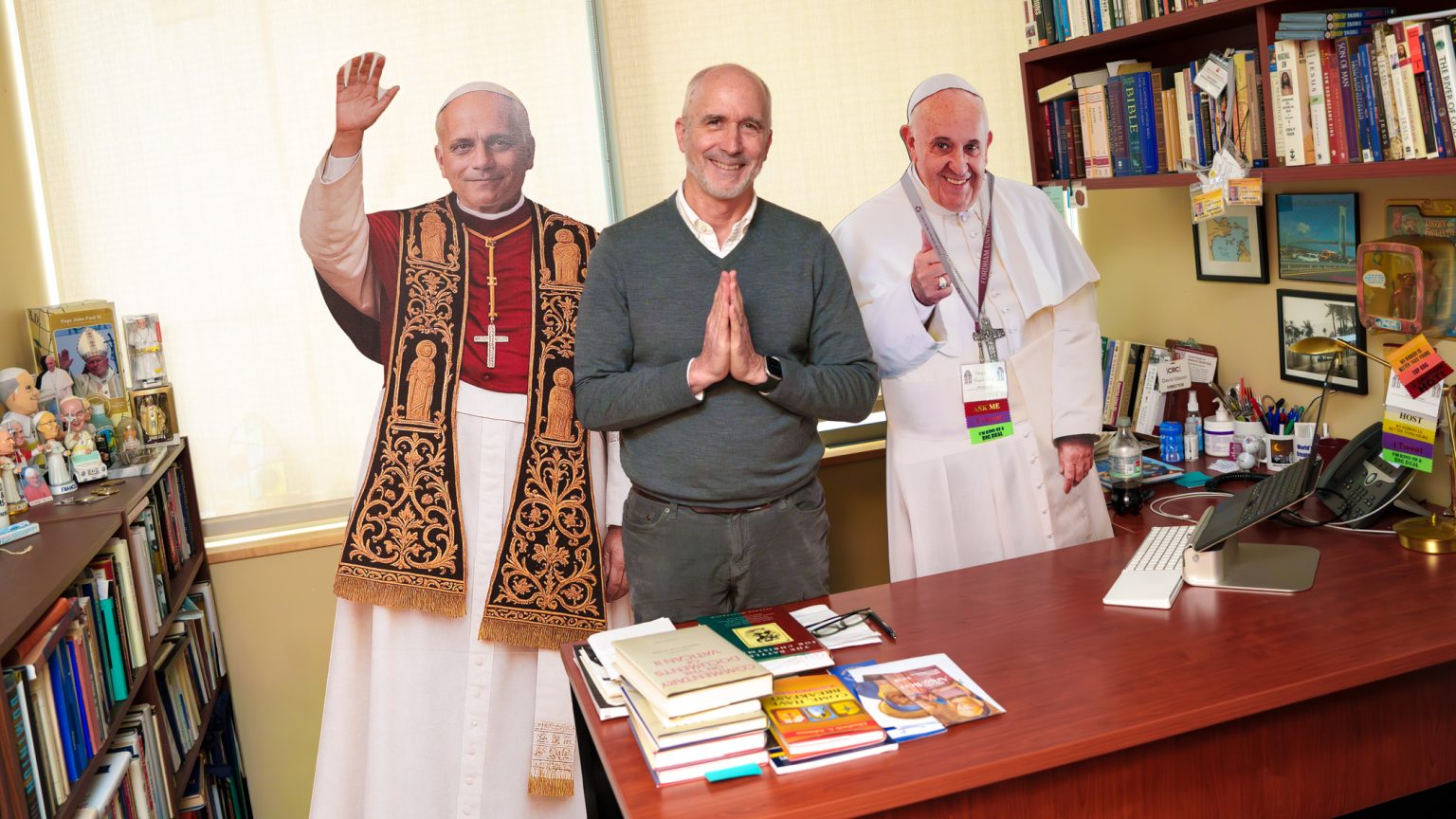 What’s on My Desk: David Gibson David Gibson stands, smiles, and holds his hands in prayer in between life-sized cardboard cutouts of Pope Leo and Pope Francis.