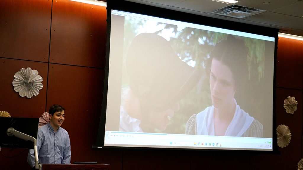 A man speaks at a podium next to a video screen.