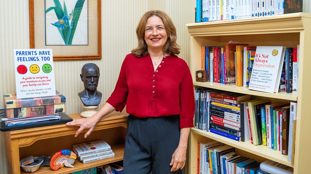 Author and Fordham grad Hilary Jacobs Hendel smiling in an office, standing next to a bookshelf and a copy of her book "Parents Have Feelings, Too."