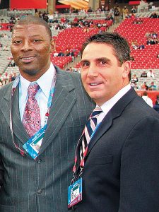 Sportscasters Carl Banks and Bob Papa stand together in suits and ties at a stadium, with red and white spectator seating visible in the background.